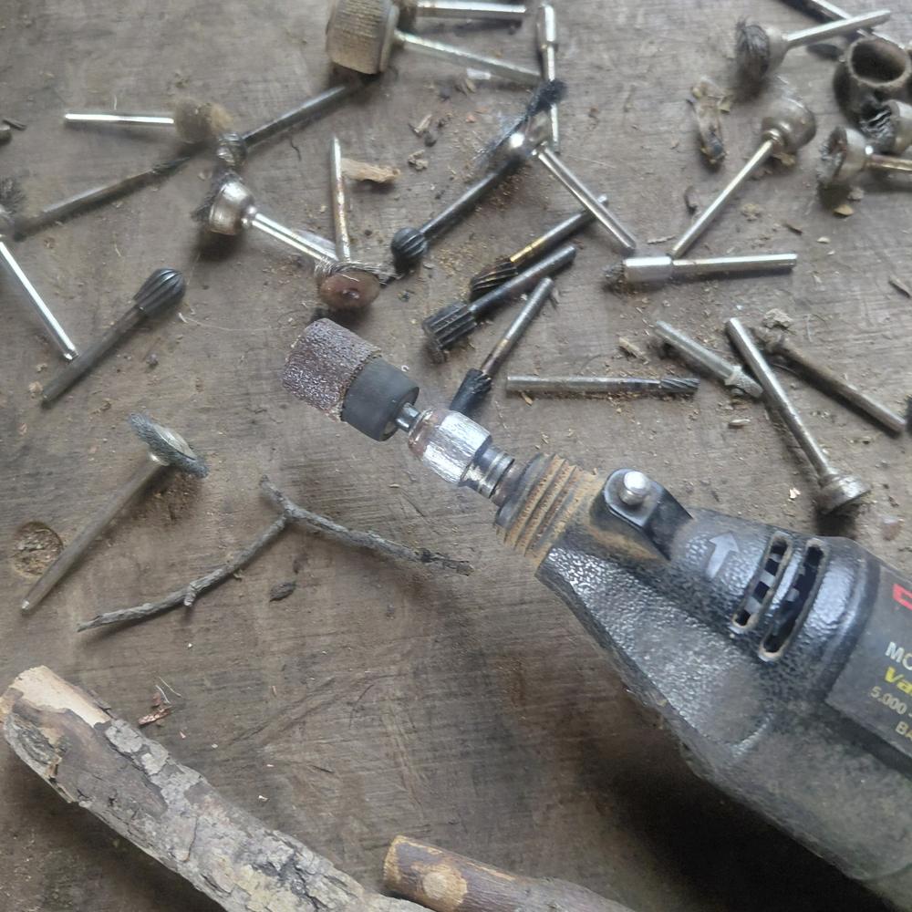 A rotary tool with a sanding drum lies on a dusty wooden bench among a scatter of burrs, wire brushes and cutting bits.