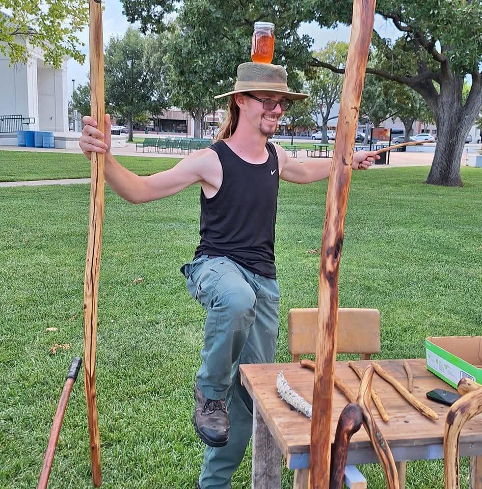 A man in a wide-brimmed hat holds two tall cedar staves at a market table in a Kansas park, a jar of honey balanced on his hat; wands and a bundle of sage are laid out on the table in front of him.