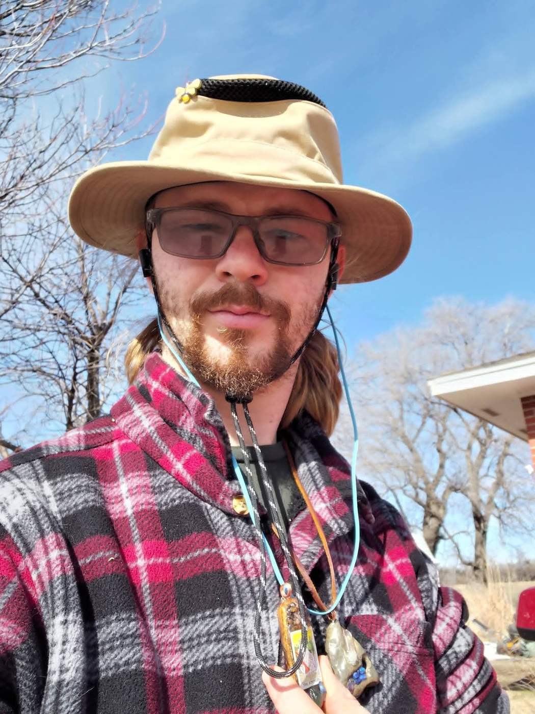 Oscar Alexander Prospero, called Sticks, in a wide hat and plaid, outdoors in Kansas, wearing two handmade pendants